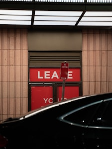 A 'No Stopping' sign is positioned in front of a glass door with red panels that display the word 'LEASE'. The scene is partially obscured by a dark vehicle, and the area is illuminated by a grid-like ceiling light.