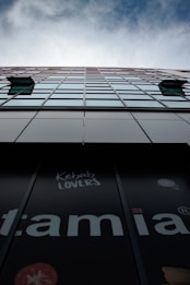 A tall mirrored building stretches into the sky, its reflective windows displaying the blue sky and clouds above. Near the base, there's signage displaying the phrase 'Kebab Lovers', with partial letters visible and elements such as cut tomatoes.