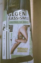 A poster displays the phrase 'GEGEN RASSISMUS' followed by the date 'FREITAG, 18. MÄRZ'. Below, there is an image of a person lying on a bed, with the text 'GERMAN CONNEC' over it. Various organizer names and logos are present, including a logo on the left side that resembles a megaphone.