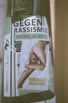 A poster displays the phrase 'GEGEN RASSISMUS' followed by the date 'FREITAG, 18. MÄRZ'. Below, there is an image of a person lying on a bed, with the text 'GERMAN CONNEC' over it. Various organizer names and logos are present, including a logo on the left side that resembles a megaphone.