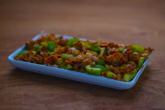 A rectangular white dish holds a flavorful stir-fry featuring vibrant green peppers and well-seasoned pieces of chicken. The dish is set on a wooden surface, showcasing a blend of textures and colors with golden-brown chicken pieces and bright green peppers.