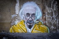 A mural on a weathered wall depicts an older man with distinctive features, including white hair and thick-rimmed glasses. He wears a yellow jacket. Mathematical and scientific equations are painted beside him, along with diagrams and chemical structures.