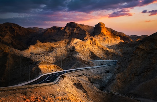 A winding road cuts through a rugged mountainous landscape bathed in warm, golden light from the setting sun. The sky is a dramatic mix of deep blues and vibrant oranges and pinks. The mountains have sharp, jagged formations with visible rocky textures.