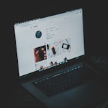 A laptop displaying an Instagram profile page, emitting a soft glow in an otherwise dark environment. The screen shows a grid of images and profile details, including followers and posts.