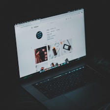 A laptop displaying an Instagram profile page, emitting a soft glow in an otherwise dark environment. The screen shows a grid of images and profile details, including followers and posts.