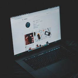 A laptop displaying an Instagram profile page, emitting a soft glow in an otherwise dark environment. The screen shows a grid of images and profile details, including followers and posts.