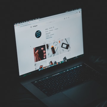 A laptop displaying an Instagram profile page, emitting a soft glow in an otherwise dark environment. The screen shows a grid of images and profile details, including followers and posts.