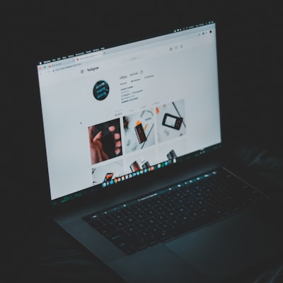 A laptop displaying an Instagram profile page, emitting a soft glow in an otherwise dark environment. The screen shows a grid of images and profile details, including followers and posts.