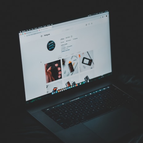 A laptop displaying an Instagram profile page, emitting a soft glow in an otherwise dark environment. The screen shows a grid of images and profile details, including followers and posts.