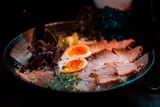 A delicious bowl of ramen features sliced pork, soft-boiled eggs with runny yolks, seaweed, mushrooms, and green onions in a rich broth, with a spoon resting on the side.