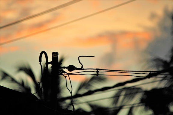 Silhouette of electrical wires and a power pole against a colorful sunset sky. The background features soft hues of orange, pink, and blue, with some palm tree outlines visible, creating a tropical atmosphere.
