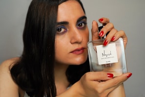 A person with long dark hair and makeup, including purple eyeshadow and red lipstick, is holding a clear perfume bottle labeled 'Nayel For Her' with both hands. The fingernails are painted red, matching the lipstick.