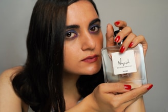 A person with long dark hair and makeup, including purple eyeshadow and red lipstick, is holding a clear perfume bottle labeled 'Nayel For Her' with both hands. The fingernails are painted red, matching the lipstick.