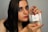 A person with long dark hair and makeup, including purple eyeshadow and red lipstick, is holding a clear perfume bottle labeled 'Nayel For Her' with both hands. The fingernails are painted red, matching the lipstick.