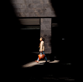 A model carrying a sleek, modern handbag while walking through a sunlit urban setting with subtle greenery.