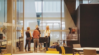 Four people are gathered around a foosball table in a modern, glass-enclosed office space. The room has large windows, contemporary furniture, and a display case against a dark wall.