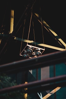 A set of four industrial lights hangs suspended from a geometric metal frame. The lights and structure are set against a dark background, with faint reflections of light visible. Metal beams create a triangular pattern, and a railing crosses the lower foreground.