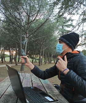 A person wearing a mask and a beanie is sitting at a wooden picnic table in a forested park, holding a handheld device. A laptop is open in front of them on the table. The trees have thick green foliage, and the setting appears calm, with a hint of sunset light filtering through.
