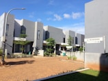 A modern residential area with minimalist architecture, featuring several two-story buildings with a gray exterior. The buildings are lined along a paved pathway, and there are some small trees and shrubs planted around, providing a touch of greenery. The sky is clear with a few clouds, indicating a sunny day. A street sign with the name Jl. Wijaksana is visible, along with a street lamp.