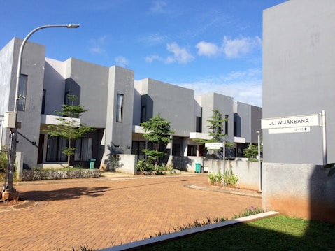 A modern residential area with minimalist architecture, featuring several two-story buildings with a gray exterior. The buildings are lined along a paved pathway, and there are some small trees and shrubs planted around, providing a touch of greenery. The sky is clear with a few clouds, indicating a sunny day. A street sign with the name Jl. Wijaksana is visible, along with a street lamp.