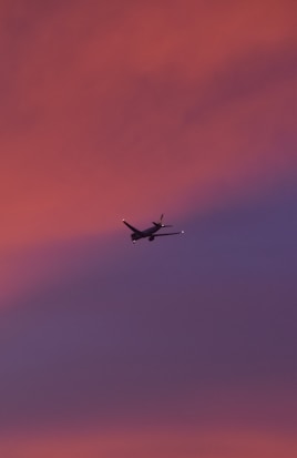 An airplane is flying across a vibrant sky during sunset or sunrise, with hues of pink, orange, and purple blending beautifully.