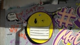 Graffiti art featuring a large yellow emoticon wearing a white face mask, painted on a gray wall. There are various colorful tags and writings in purple, red, and orange, including a large hash symbol and the word 'SATO' above the emoticon. Additionally, a speech bubble with text is located on the left side.