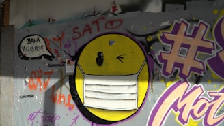 Graffiti art featuring a large yellow emoticon wearing a white face mask, painted on a gray wall. There are various colorful tags and writings in purple, red, and orange, including a large hash symbol and the word 'SATO' above the emoticon. Additionally, a speech bubble with text is located on the left side.