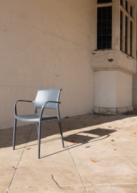 A simple grey plastic chair sits on a tiled outdoor patio against an old, weathered wall. The wall has an architectural feature with large, multi-paned windows. Autumn leaves are scattered across the patio, and the chair casts a long shadow in the sunlight.