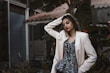 A fashionably dressed person stands in a relaxed pose, with one hand resting on their head. They wear a light-colored blazer over a patterned top and vibrant lipstick. Sunglasses dangle from the blazer. The background features lush greenery and parts of a building structure, giving an urban, natural juxtaposition.