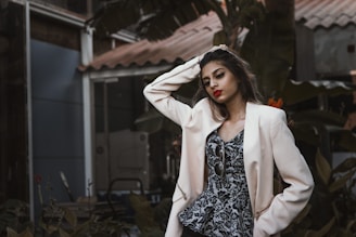 A fashionably dressed person stands in a relaxed pose, with one hand resting on their head. They wear a light-colored blazer over a patterned top and vibrant lipstick. Sunglasses dangle from the blazer. The background features lush greenery and parts of a building structure, giving an urban, natural juxtaposition.