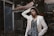 A fashionably dressed person stands in a relaxed pose, with one hand resting on their head. They wear a light-colored blazer over a patterned top and vibrant lipstick. Sunglasses dangle from the blazer. The background features lush greenery and parts of a building structure, giving an urban, natural juxtaposition.