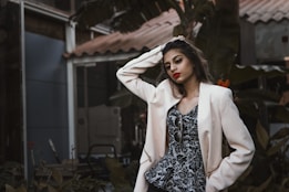 A fashionably dressed person stands in a relaxed pose, with one hand resting on their head. They wear a light-colored blazer over a patterned top and vibrant lipstick. Sunglasses dangle from the blazer. The background features lush greenery and parts of a building structure, giving an urban, natural juxtaposition.
