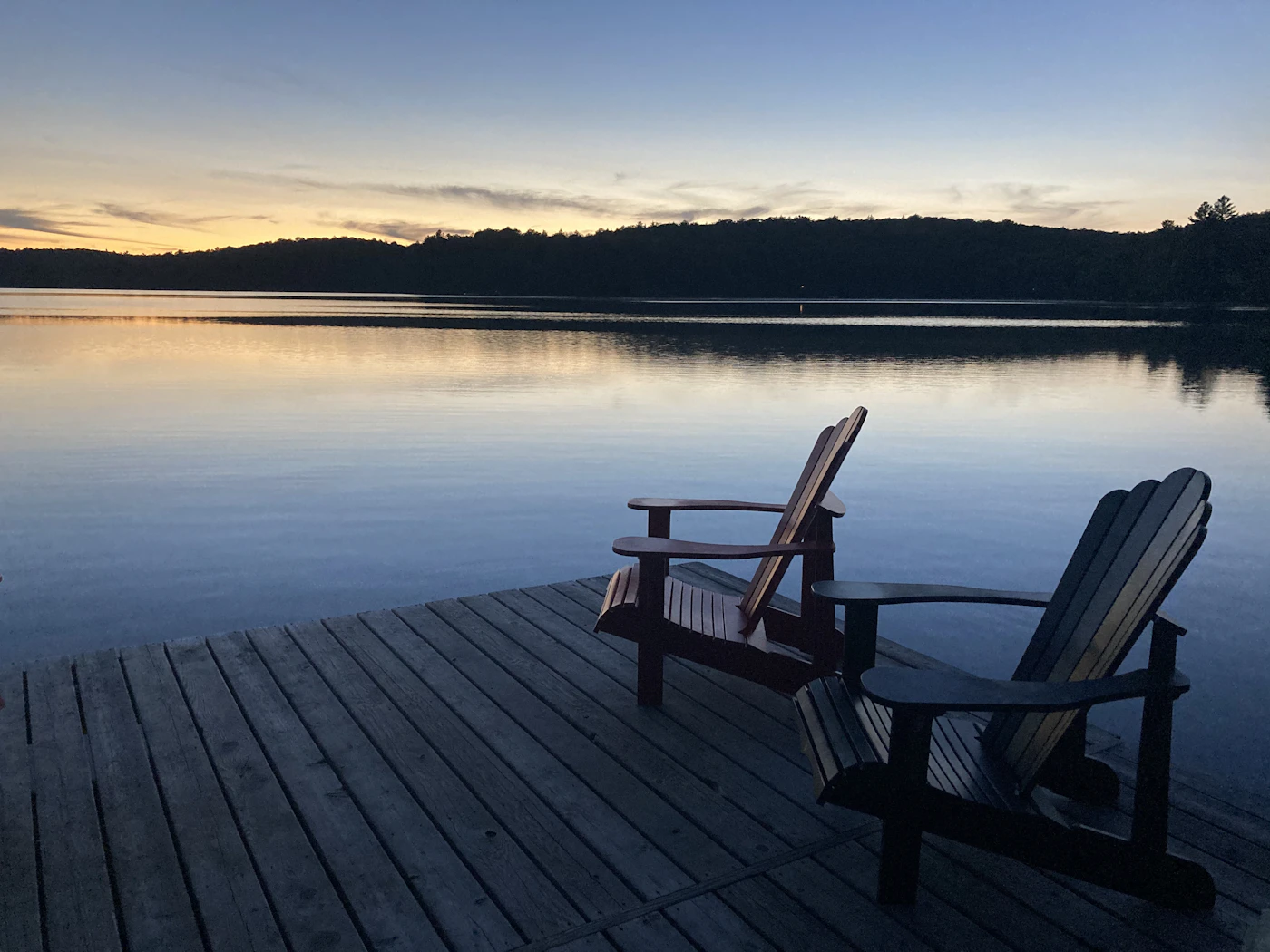 Two wooden Muskoka chairs on a dock facing a calm Ontario lake at dusk, the silhouette of forested hills across the water under a soft sky