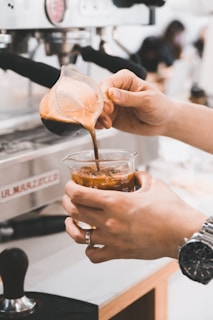 A person is pouring freshly brewed espresso from a glass decanter into a beaker filled with ice. The scene is set at a coffee machine in a cafe, emphasizing the art of making iced coffee. The person's wrist is adorned with a watch, adding a touch of style to the process.