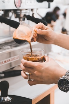 A person is pouring freshly brewed espresso from a glass decanter into a beaker filled with ice. The scene is set at a coffee machine in a cafe, emphasizing the art of making iced coffee. The person's wrist is adorned with a watch, adding a touch of style to the process.