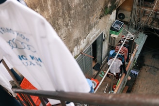 The photo captures a small balcony area with a clothesline and several pieces of clothing hanging to dry. The balcony wall appears aged and dirty, with visible peeling paint. Various household items like a basket, buckets, and sandals are scattered around. An air conditioning unit is mounted on the wall under a series of tangled wires. The overall scene conveys a sense of urban residential life.