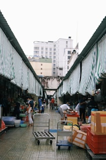 A bustling outdoor market with striped awnings where several people are engaged in various activities. There are carts, boxes, and baskets on the ground, while customers and vendors interact by the stalls. The market is flanked by buildings under an overcast sky.