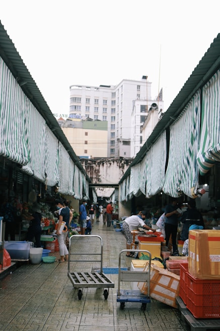 A bustling outdoor market with striped awnings where several people are engaged in various activities. There are carts, boxes, and baskets on the ground, while customers and vendors interact by the stalls. The market is flanked by buildings under an overcast sky.