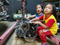 Two young children are sitting in a workshop environment, interacting with a car engine placed on the floor. One child is wearing a blue shirt, and the other is in a yellow shirt with red sleeves. Various mechanical tools and equipment are visible in the background.