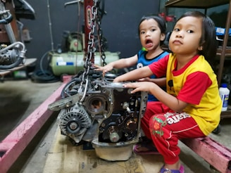 Two young children are sitting in a workshop environment, interacting with a car engine placed on the floor. One child is wearing a blue shirt, and the other is in a yellow shirt with red sleeves. Various mechanical tools and equipment are visible in the background.