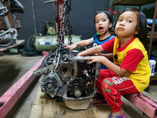 Two young children are sitting in a workshop environment, interacting with a car engine placed on the floor. One child is wearing a blue shirt, and the other is in a yellow shirt with red sleeves. Various mechanical tools and equipment are visible in the background.