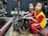 Two young children are sitting in a workshop environment, interacting with a car engine placed on the floor. One child is wearing a blue shirt, and the other is in a yellow shirt with red sleeves. Various mechanical tools and equipment are visible in the background.
