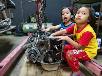 Two young children are sitting in a workshop environment, interacting with a car engine placed on the floor. One child is wearing a blue shirt, and the other is in a yellow shirt with red sleeves. Various mechanical tools and equipment are visible in the background.