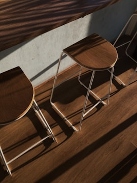 Wooden stools with metal frames are arranged along a bar table. Sunlight creates sharp shadows and highlights the natural grain of the wood. The floor and table also have a similar brown wood finish, enhancing the warm and cozy atmosphere.