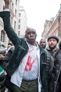 A man stands among a group, raising his fist assertively. He wears glasses, a dark green jacket, and a white shirt stained with red, resembling blood splatter. The crowd behind him appears to be in an urban setting, with tall buildings visible in the background.