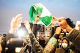 A group of people gather together, raising their arms in the air in a show of solidarity, with one person prominently holding up a green and white flag amid the crowd. The scene is vibrant and dynamic, capturing the energy and enthusiasm of the group, with some individuals wearing masks.
