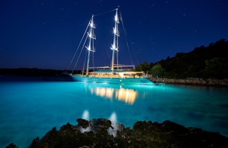 A large, illuminated yacht is anchored in a serene bay with calm waters. The sky is dark, dotted with stars, and the yacht's lights cast a vibrant reflection on the clear, turquoise water. The shore is outlined with silhouettes of trees and rocky formations.