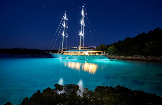 A large, illuminated yacht is anchored in a serene bay with calm waters. The sky is dark, dotted with stars, and the yacht's lights cast a vibrant reflection on the clear, turquoise water. The shore is outlined with silhouettes of trees and rocky formations.