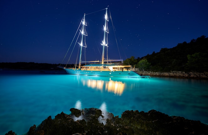 A large, illuminated yacht is anchored in a serene bay with calm waters. The sky is dark, dotted with stars, and the yacht's lights cast a vibrant reflection on the clear, turquoise water. The shore is outlined with silhouettes of trees and rocky formations.
