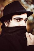 A person is wearing a black fedora and a coat with the collar pulled up, covering most of their lower face. The focus is on their eyes, which convey a mysterious or secretive expression. The background features a soft bokeh effect with warm tones of brown and green.